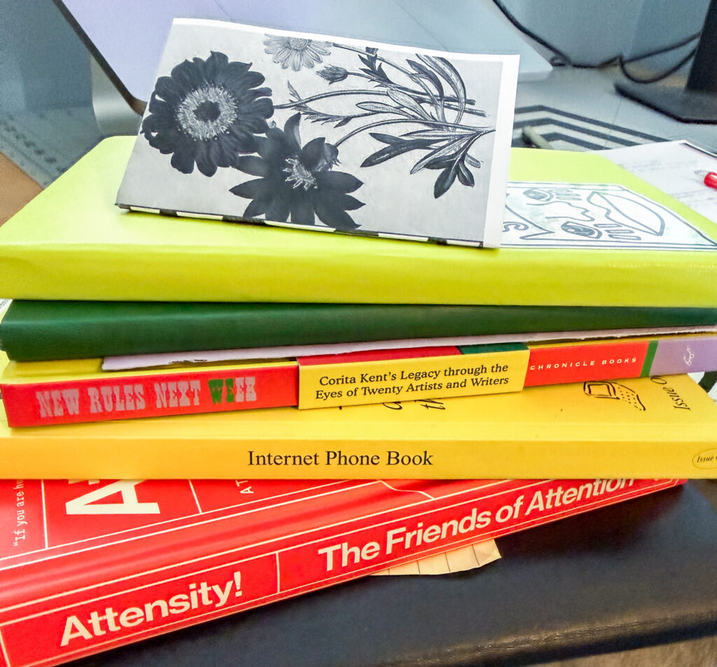 a stack of books. from bottom, Attensity!, Internet Phone Book, New Rules Next Week, two Moleskine notebooks, and a mini folded zine with black and white florals.